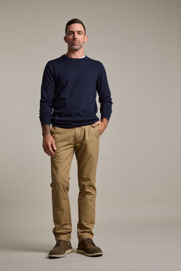 A man with short dark hair and a neutral expression stands in a beige studio, wearing a navy sweater, Barkers Dundas Chino relaxed fit trousers in lightweight cotton twill, and brown shoes. One hand is in his pocket.