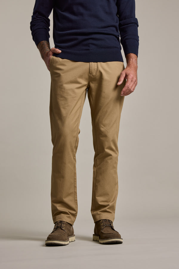 A person models Barkers Dundas Chino trousers in tan khaki, paired with a navy sweater and brown shoes, standing with one hand in their pocket against a neutral background; only the lower body is visible.