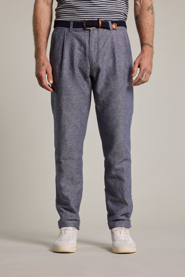 A person, visible from shoulders down, wears Barkers Newman Linen Pants in blue with a dark belt, a tucked-in striped shirt, and white sneakers, standing against a plain light background.