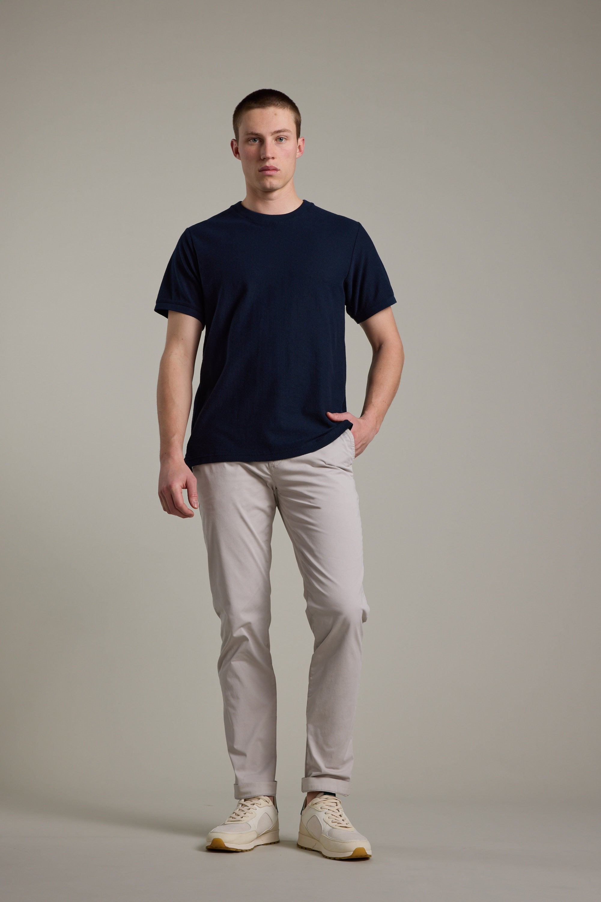 A man stands against a plain background, wearing a navy t-shirt, Barkers Kingly Chino Pant in light beige, and white sneakers. With one hand in his pocket and a neutral expression, he showcases these summer-ready chinos.