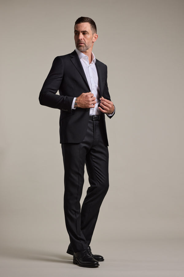 A man with short hair and a beard stands against a plain background, wearing the Barkers Mayfair ZQrx Suit Jacket over a white dress shirt and black dress shoes. He adjusts his jacket with one hand and gazes to the side.