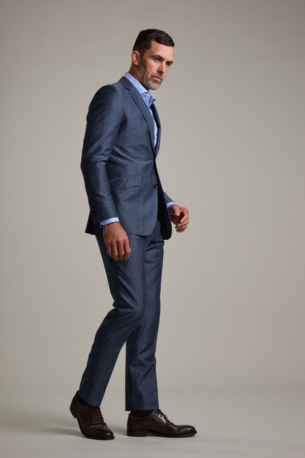A man stands sideways against a plain light background, wearing Barkers' Charles Texture Suit Trouser in steel grey, paired with a light blue dress shirt and brown dress shoes, looking slightly toward the camera.