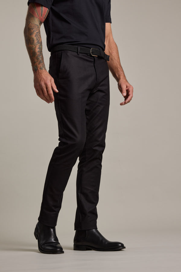A man stands against a plain background wearing black boots, a black T-shirt, and Barkers Lyndon Cotton Pants with a belt. His tattooed left arm is relaxed at his side; his head is out of frame.