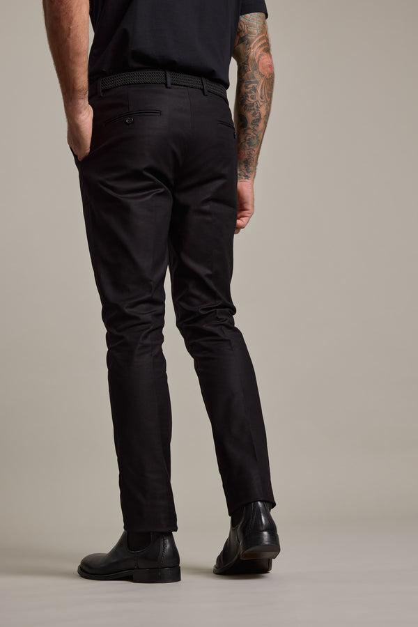 A man in smart casual style, wearing Barkers Lyndon Cotton Pants, a black belt, and black shoes stands with his tattooed right arm by his side, facing a plain light background.