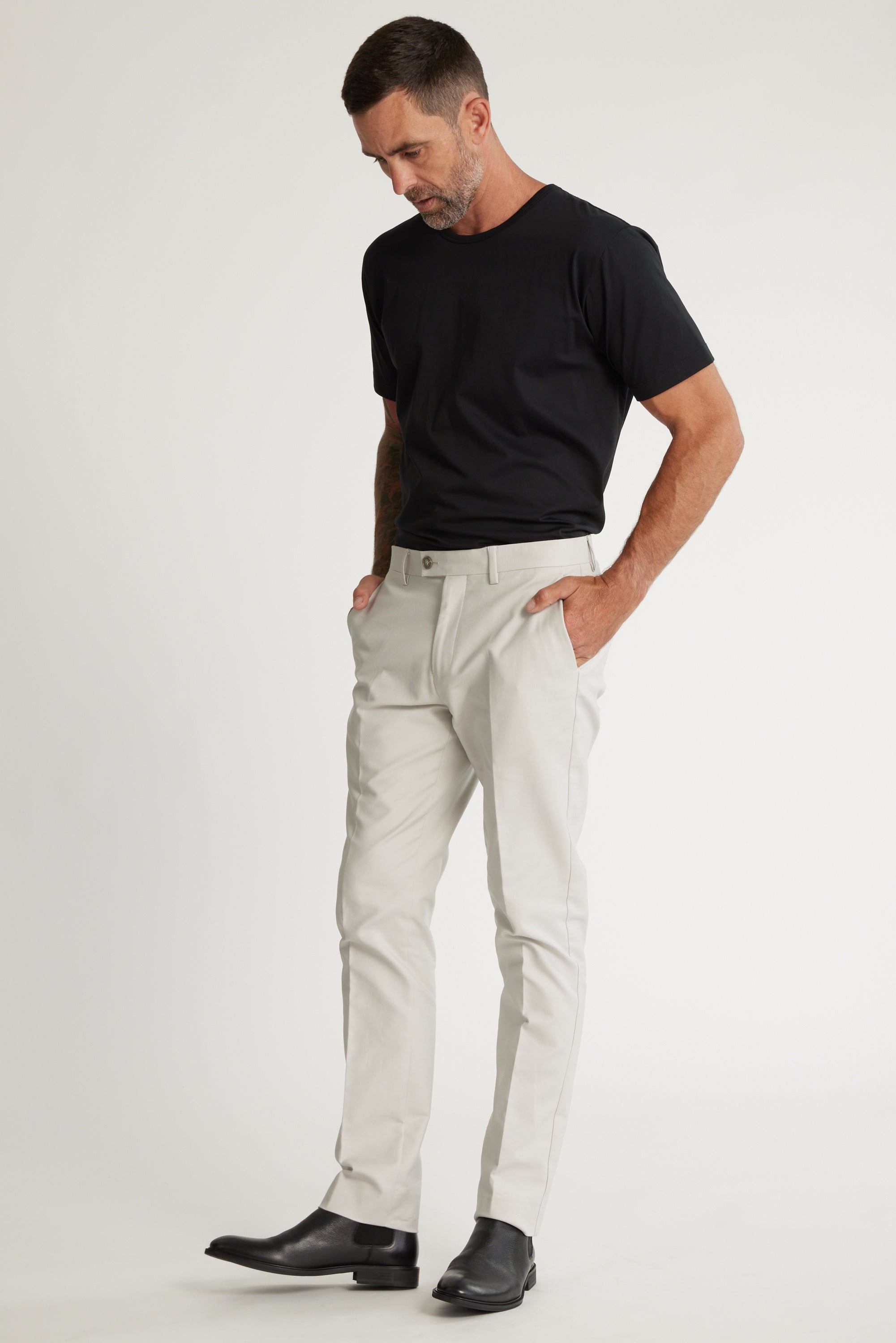 A man models Barkers' Lyndon Cotton Pants, paired with a black t-shirt and shoes. With short hair and a beard, he looks down with hands in pockets, showcasing an effortlessly smart casual style against a white background.