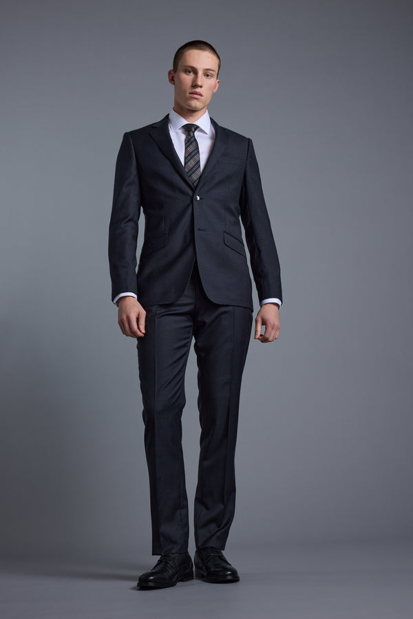 A man stands against a gray background, wearing the Barkers Mayfair ZQ Suit Jacket made from Merino Wool, paired with a white shirt, striped tie, and black dress shoes. He faces forward with a neutral expression and relaxed arms at his sides.