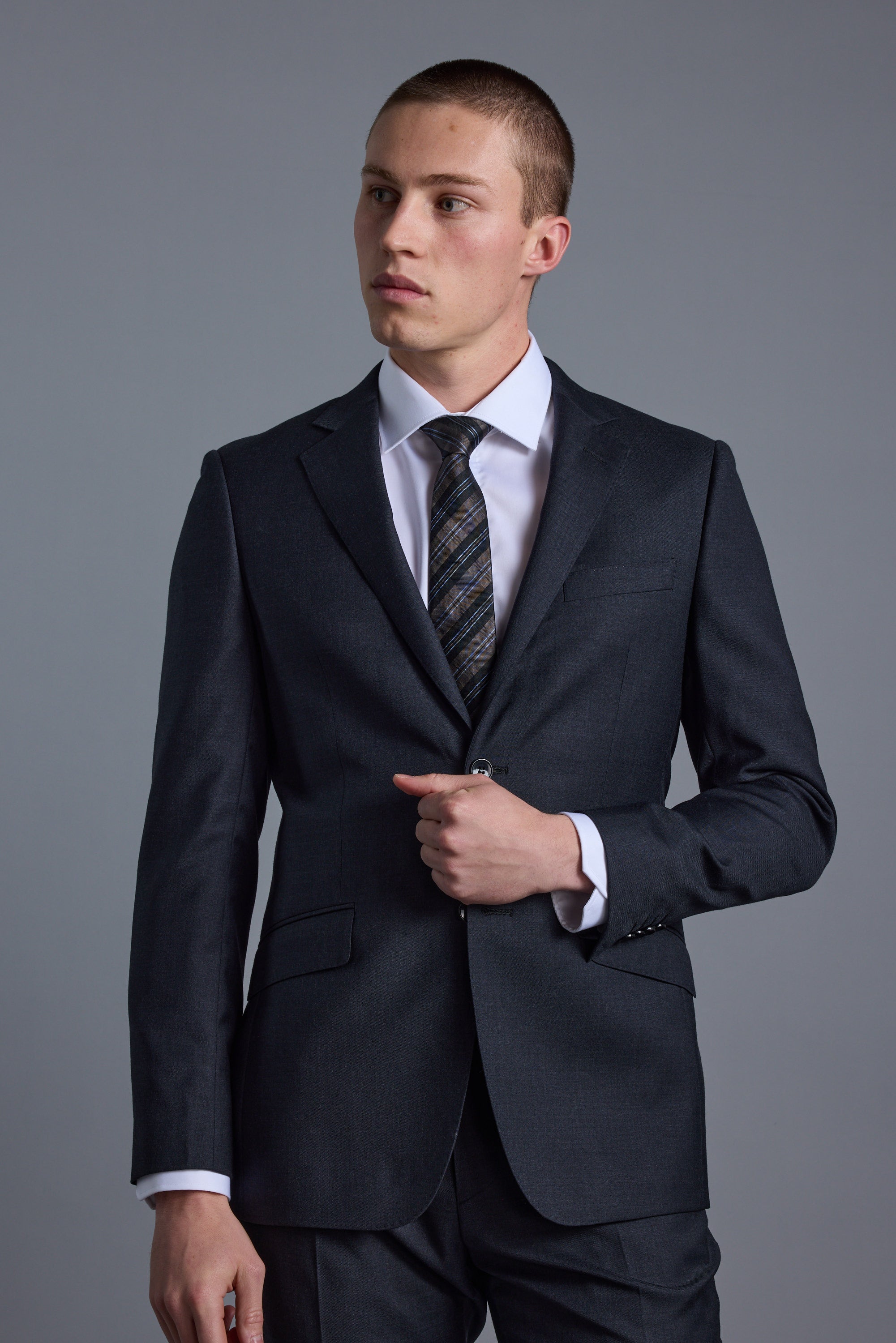 A young man in the Barkers Mayfair ZQ Suit Jacket, made from merino wool, stands against a plain gray background, gazing slightly to the side with a neutral expression.