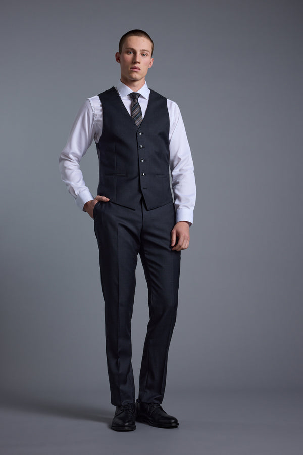 A man stands against a gray background wearing Barkers Mayfair ZQrx Suit Trousers with a matching dark jacket, white dress shirt, patterned tie, and black dress shoes, one hand in his pocket and a neutral expression.