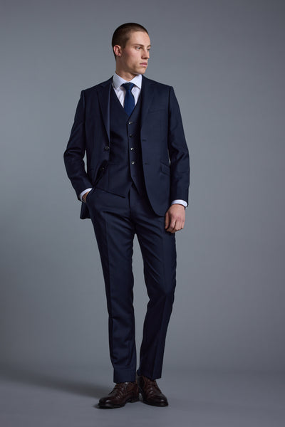 Man in Barkers Mayfair ZQrx Suit Trouser, white shirt, and blue tie stands before a gray background.