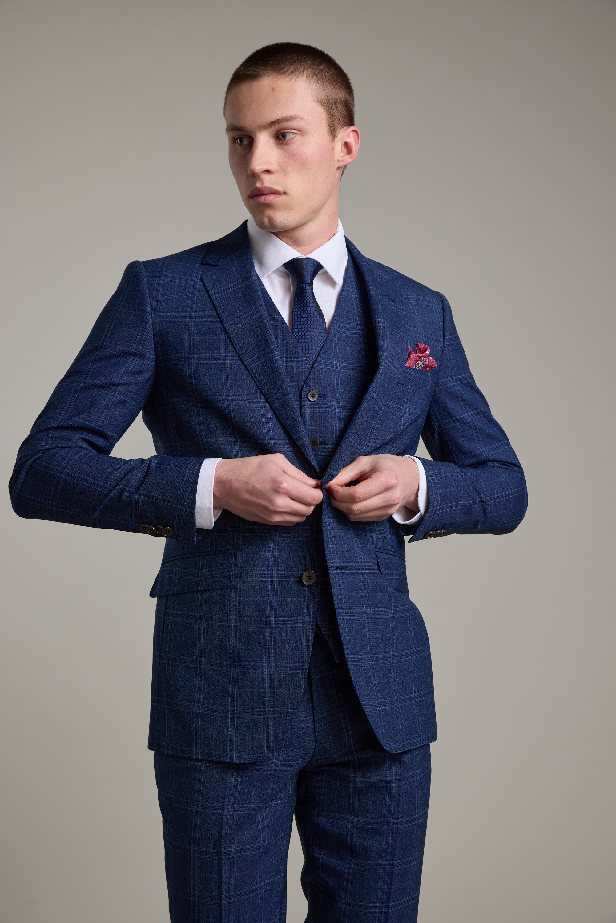 A young man in the Barkers Leonard Check Suit Jacket, styled with a white shirt, dark tie, and red pocket square, stands against a plain light background. He has short hair and a serious expression as he adjusts his jacket button.