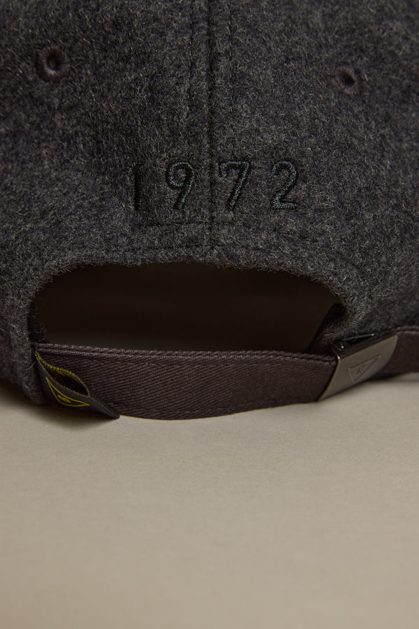 Close-up of the back of the Barkers Darcy Woollen Cap in dark gray wool, featuring "1972" embroidery above a brown adjustable strap with metal clasp. Cap rests on a light beige surface.