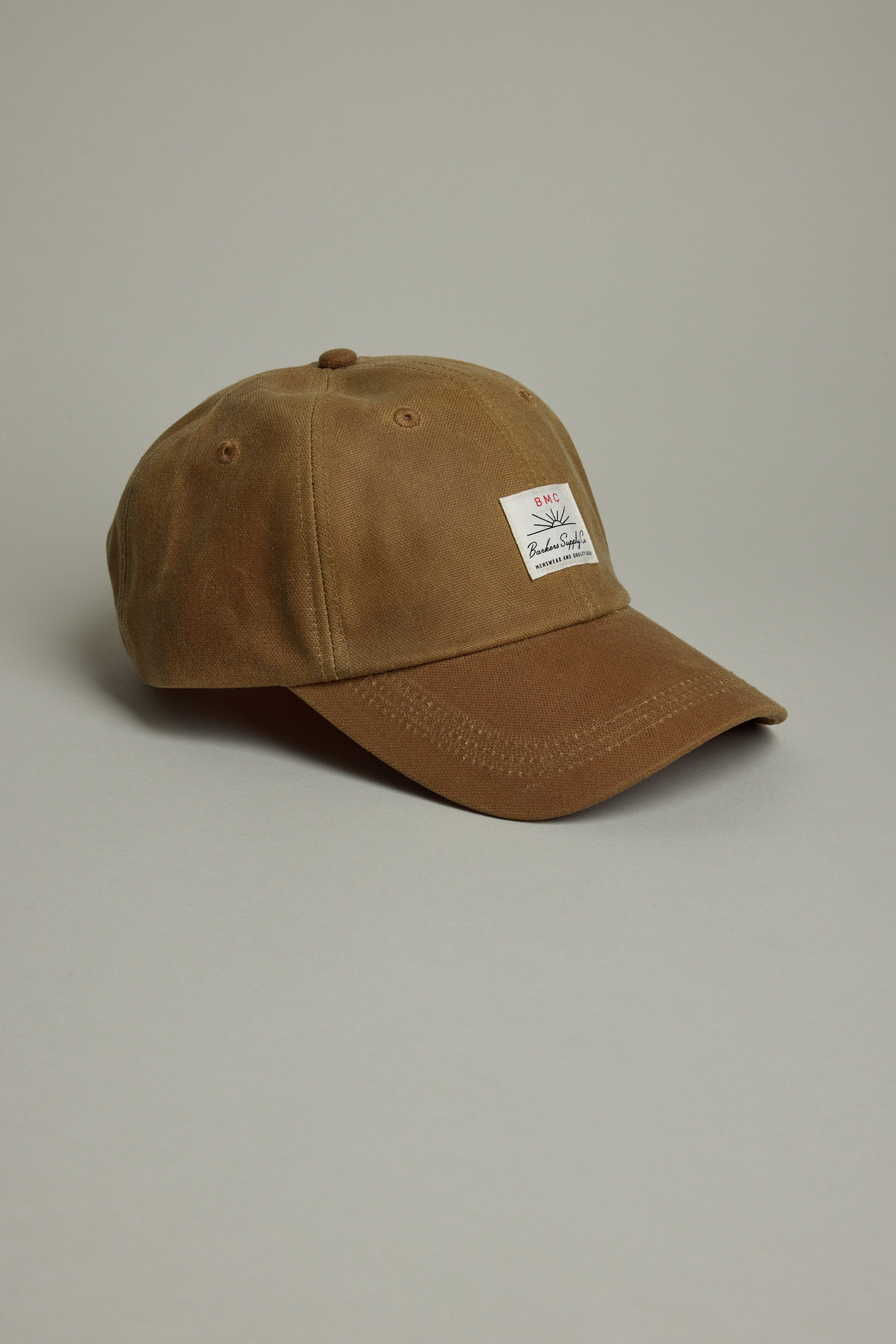 The Barkers Benson Waxed Cotton Cap features brown waxed cotton, a curved brim, and a white rectangular front patch. It offers an adjustable fit and is shown against a plain light gray background.