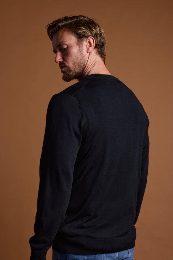 A man with light brown hair and a beard stands with his back to the camera, wearing a black Barkers Skyland ZQrx Merino Crew sweater and blue jeans against a plain brown background while looking over his shoulder.