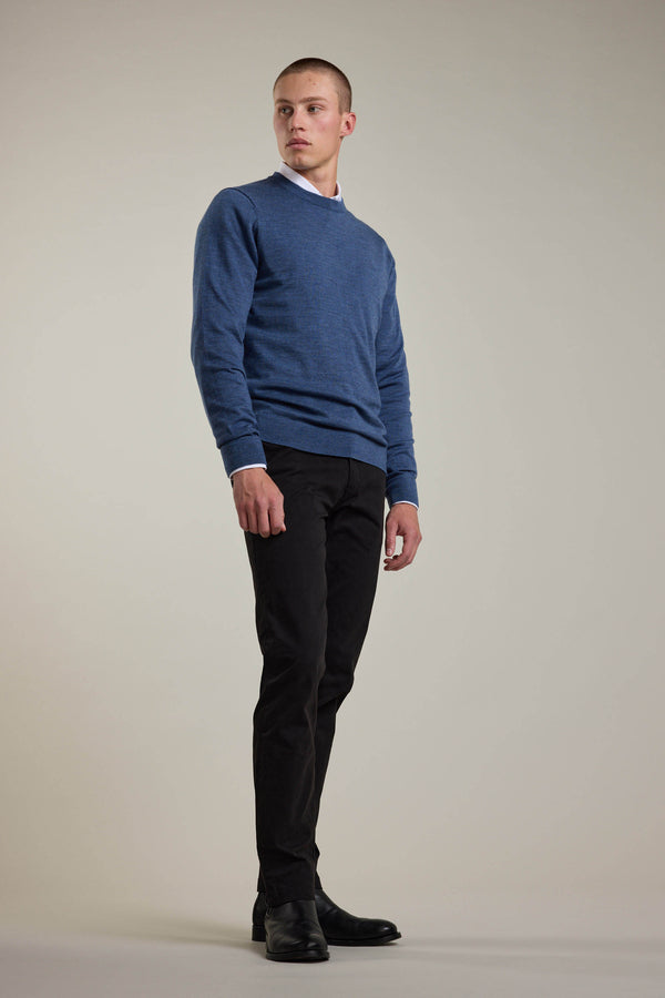 A man stands against a light gray background wearing a blue Barkers Skyland ZQrx Merino Crew sweater made from ethically sourced New Zealand merino wool, paired with black pants and shoes, gazing slightly to the side with a neutral expression.