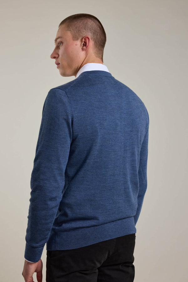 A person in a blue Barkers Skyland ZQrx Merino Crew sweater, crafted from ethically sourced merino, stands mostly with their back to the camera, glancing over their left shoulder against a plain light background.