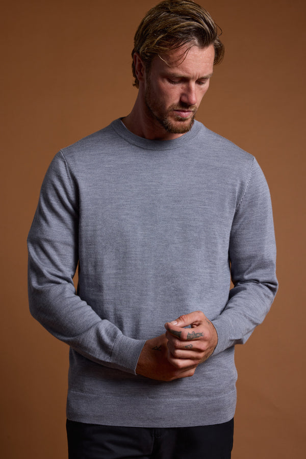 A man with short, light brown hair and a beard wears the Barkers Skyland ZQrx Merino Crew, made from sustainable New Zealand merino wool. He stands against a brown background, looking down with his hands clasped.