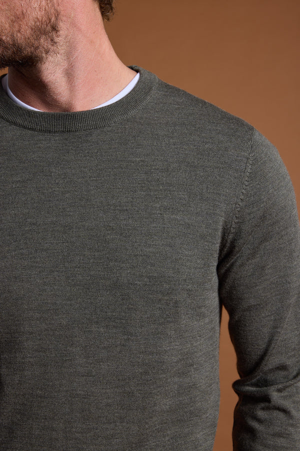 A close-up shows a person wearing the Barkers Skyland ZQrx Merino Crew in dark grey over a white t-shirt, set against a brown background. Only their lower face and neck are visible, with the head partially out of frame.