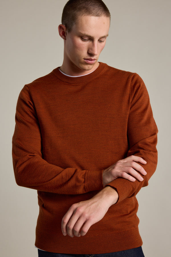 A man wearing the Barkers Skyland ZQrx Merino Crew, a rust-colored sustainable sweater, adjusts his sleeve while standing against a plain, light background.
