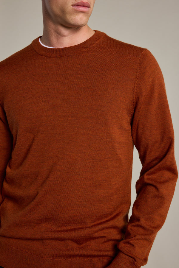 A person models the Barkers Skyland ZQrx Merino Crew, a rust-colored, long-sleeve sustainable knit sweater, against a plain beige background, showing only their lower face and upper torso.