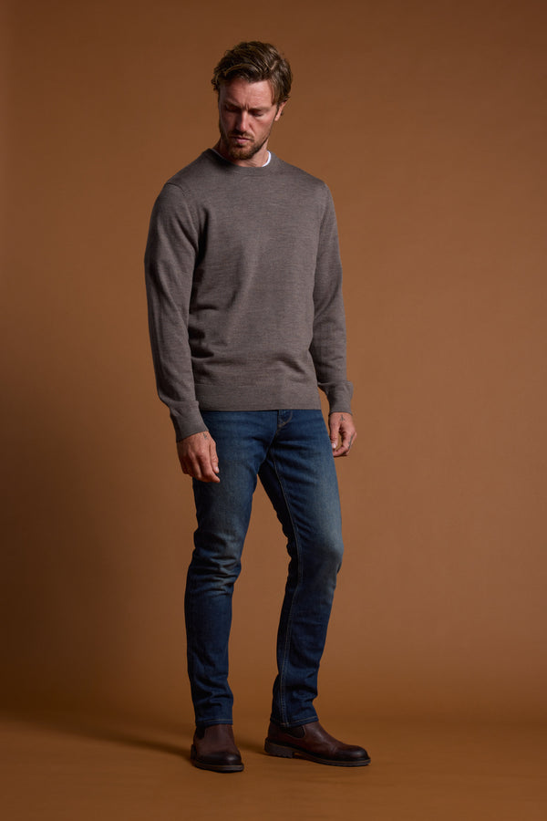 A man stands against a brown backdrop wearing the Barkers Skyland ZQrx Merino Crew, blue jeans, and brown shoes. He looks slightly downward with a neutral expression—showcasing thoughtful, sustainable wardrobe choices.