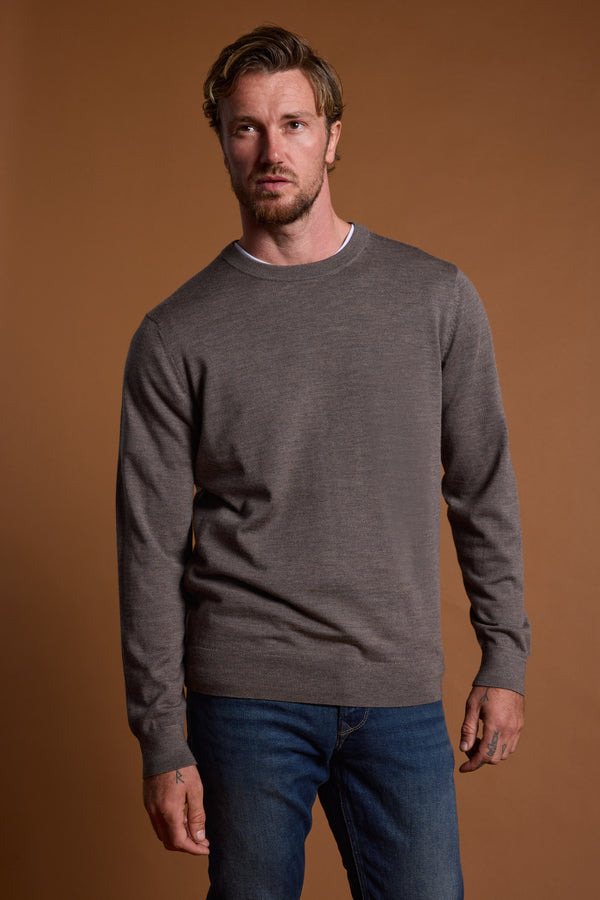 A man with short, wavy hair wears a Barkers Skyland ZQrx Merino Crew sweater in taupe over a white shirt and blue jeans, standing against a plain brown background.