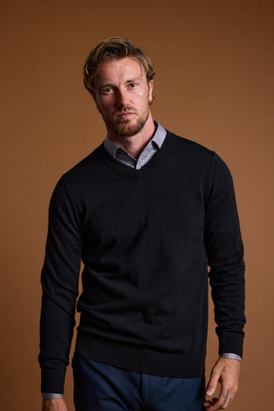 A man with light brown hair and a beard stands against a plain brown background, wearing the Barkers Skyland ZQrx Merino Vee black sweater over a light collared shirt, looking directly at the camera with a neutral expression.