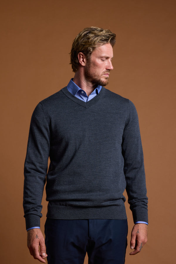 A blond man with a beard wears the Barkers Skyland ZQrx Merino Vee, a blue-collared shirt under a dark gray V-neck sweater made from breathable New Zealand merino wool, standing against a plain brown background and looking left.