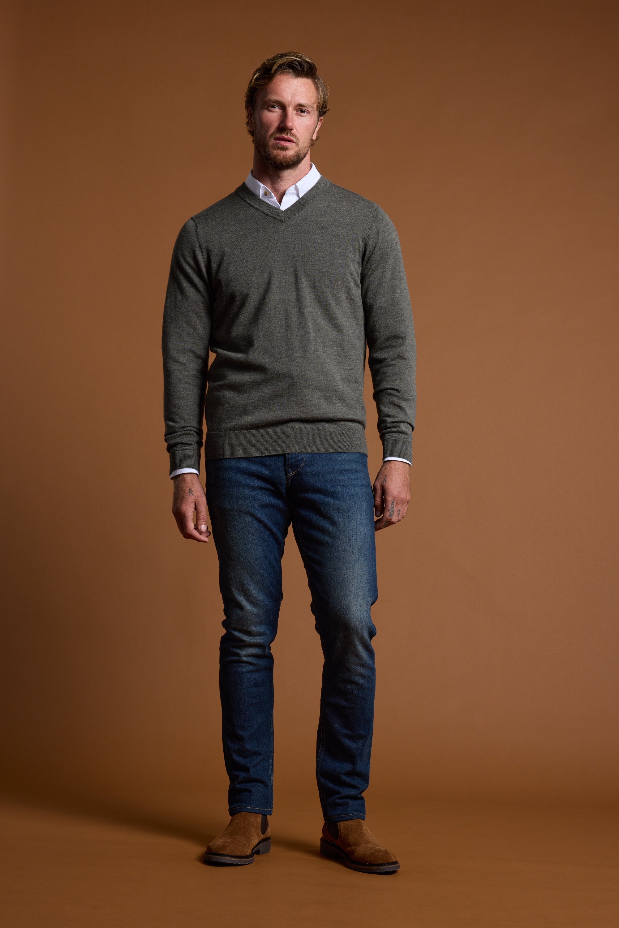A man with short blond hair and a beard stands against a brown background, wearing the Barkers Skyland ZQrx Merino Vee sweater in gray over a white shirt, with blue jeans and brown suede shoes. He has a neutral expression.