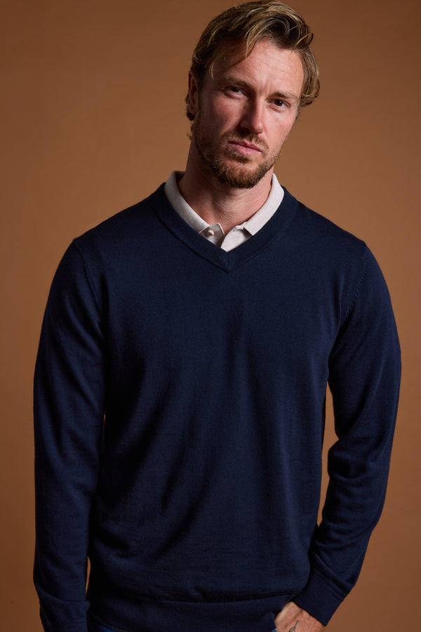 A man with light brown hair and a short beard stands against a brown background, wearing the Barkers Skyland ZQrx Merino Vee— a navy V-neck sweater made from New Zealand merino wool—over a light collared shirt. He looks neutrally at the camera.