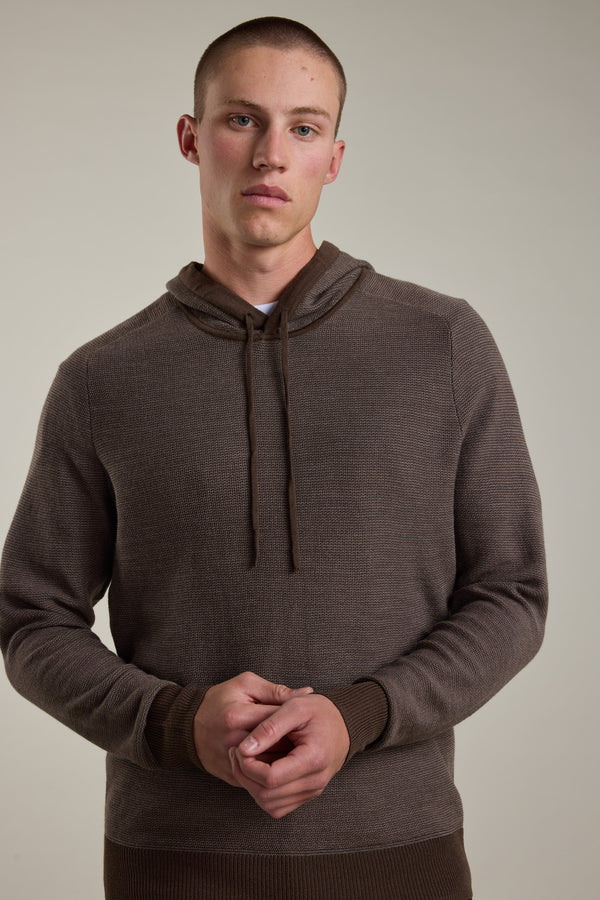 A person stands against a plain background wearing the Barkers Roxbury Merino Hoody in brown, hands loosely clasped at their waist and a neutral expression, highlighting this lightweight sweater’s effortless style.