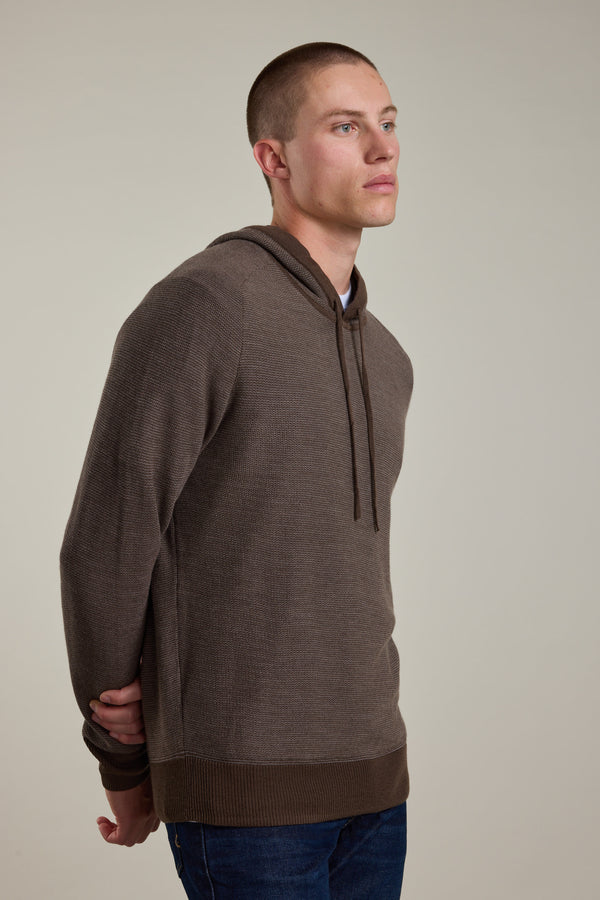 A young man with short hair wears the Barkers Roxbury Merino Hoody in brown and blue jeans, standing with his hands behind his back and gazing slightly to the side against a plain light background.