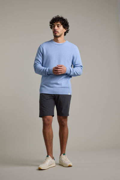 A man with curly hair stands against a plain background wearing Barkers Hutchinson Walkshort in dark gray, a light blue sweater, and white sneakers. He looks slightly to the side with his hands loosely clasped.