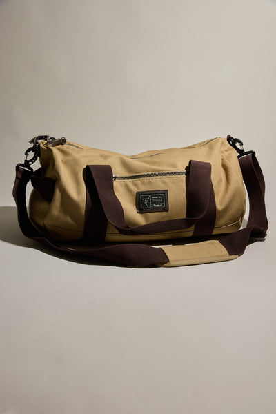 The Barkers Canvas Duffle Bag, made of tan cotton canvas with dark brown handles and a shoulder strap, features a front zipper pocket and black patch label—an ideal travel companion from Barkers.
