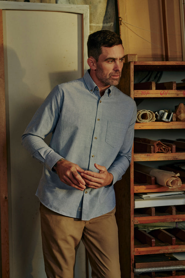 A man wears the Barkers Thatcher Oxford Shirt in light blue, crafted from responsibly sourced cotton, paired with khaki pants. He stands in a workshop with shelves of tools behind him, looking to the side with hands clasped.