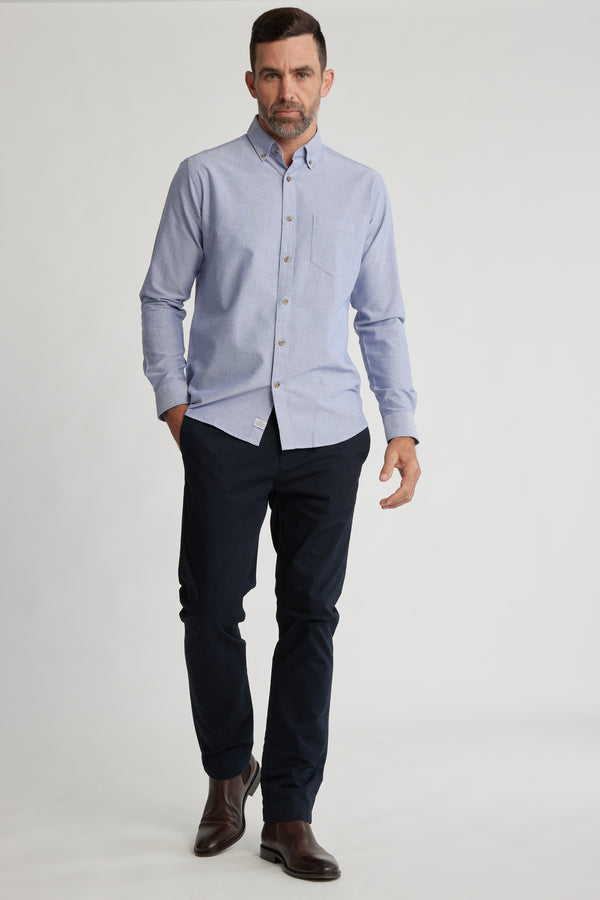 A man with short dark hair and a beard wears the Barkers Thatcher Oxford Shirt in light blue, paired with dark navy pants and brown boots, walking confidently against a plain white background.