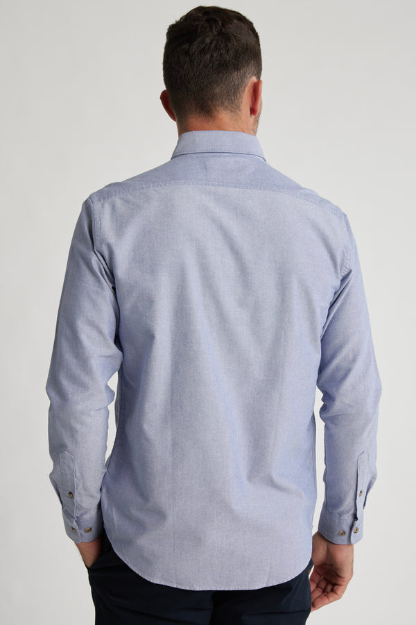 A man faces away from the camera, wearing the Barkers Thatcher Oxford Shirt in light blue—crafted from responsibly sourced cotton—with dark pants against a plain, light background.