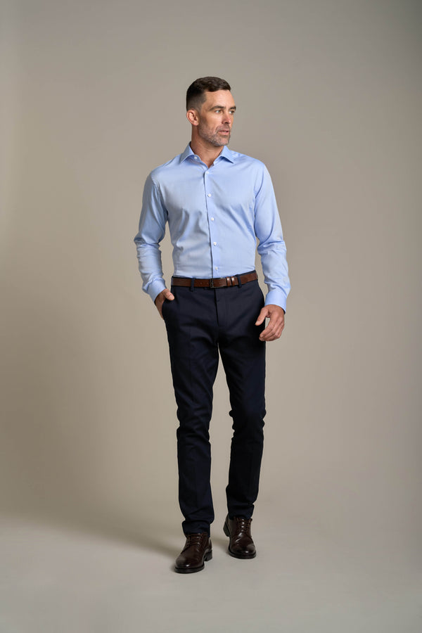 A man in a Barkers Tribeca Reg Cuff Shirt—tailored fit, light blue, wrinkle-free—paired with dark pants and brown shoes stands with one hand in his pocket, looking to the side against a plain light gray background.