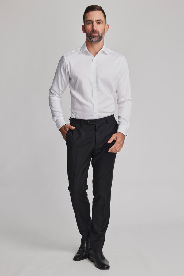 A man with short dark hair and a beard stands against a plain background, wearing the Barkers Tribeca French Cuff Shirt, black dress pants, and black shoes. His left hand is in his pocket while his right rests at his side.