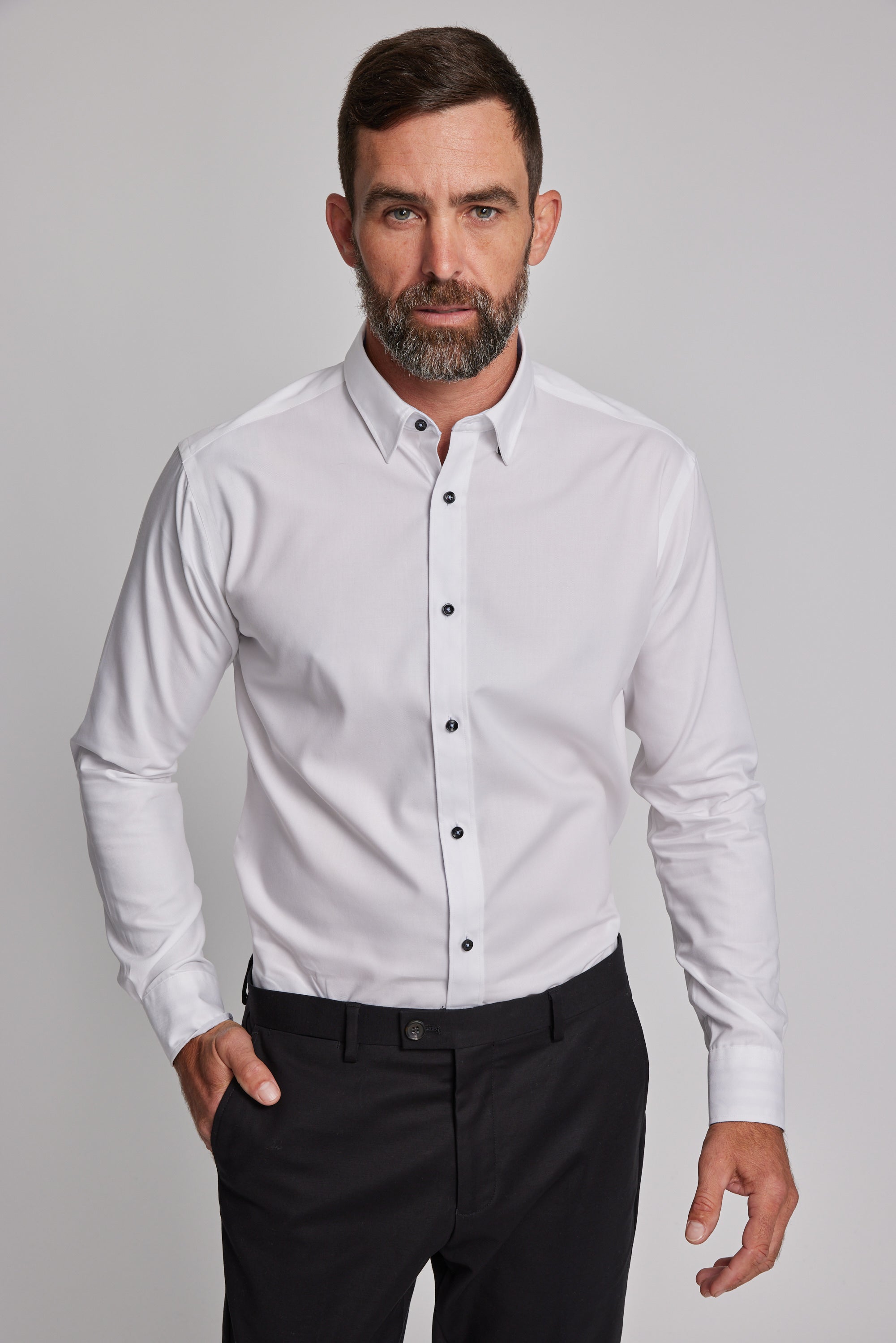 A man with short brown hair and a beard wears the Barkers Verona Shirt, a tailored fit, long sleeve white business shirt in 100% cotton, paired with black pants, standing against a plain light gray background.