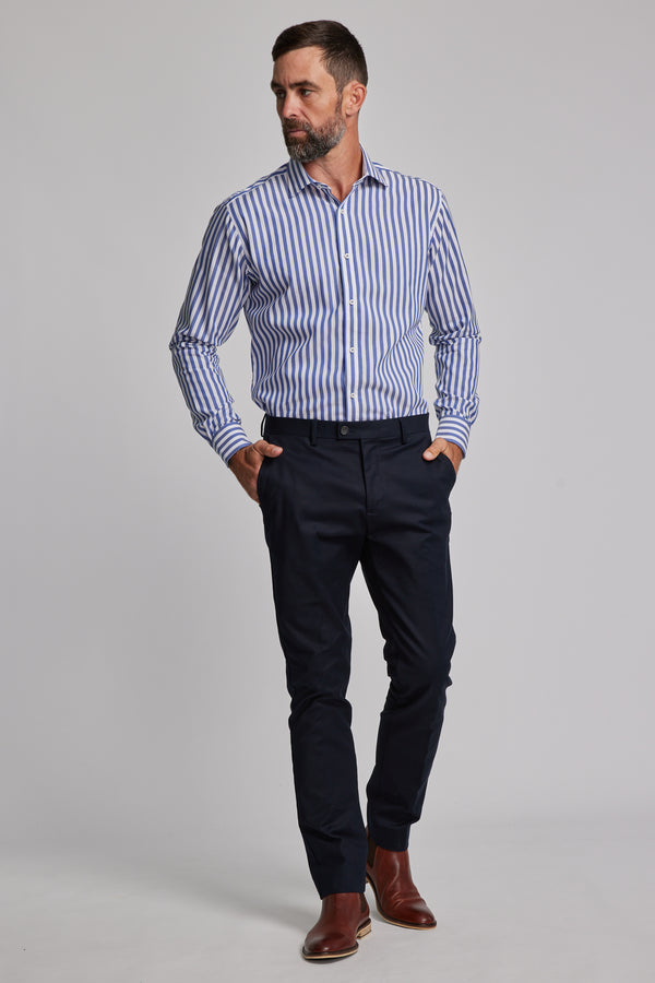 A man stands against a plain background wearing the Barkers Midway Shirt, a tailored fit blue striped business shirt, paired with dark blue pants and brown dress shoes, his hands in his pockets.