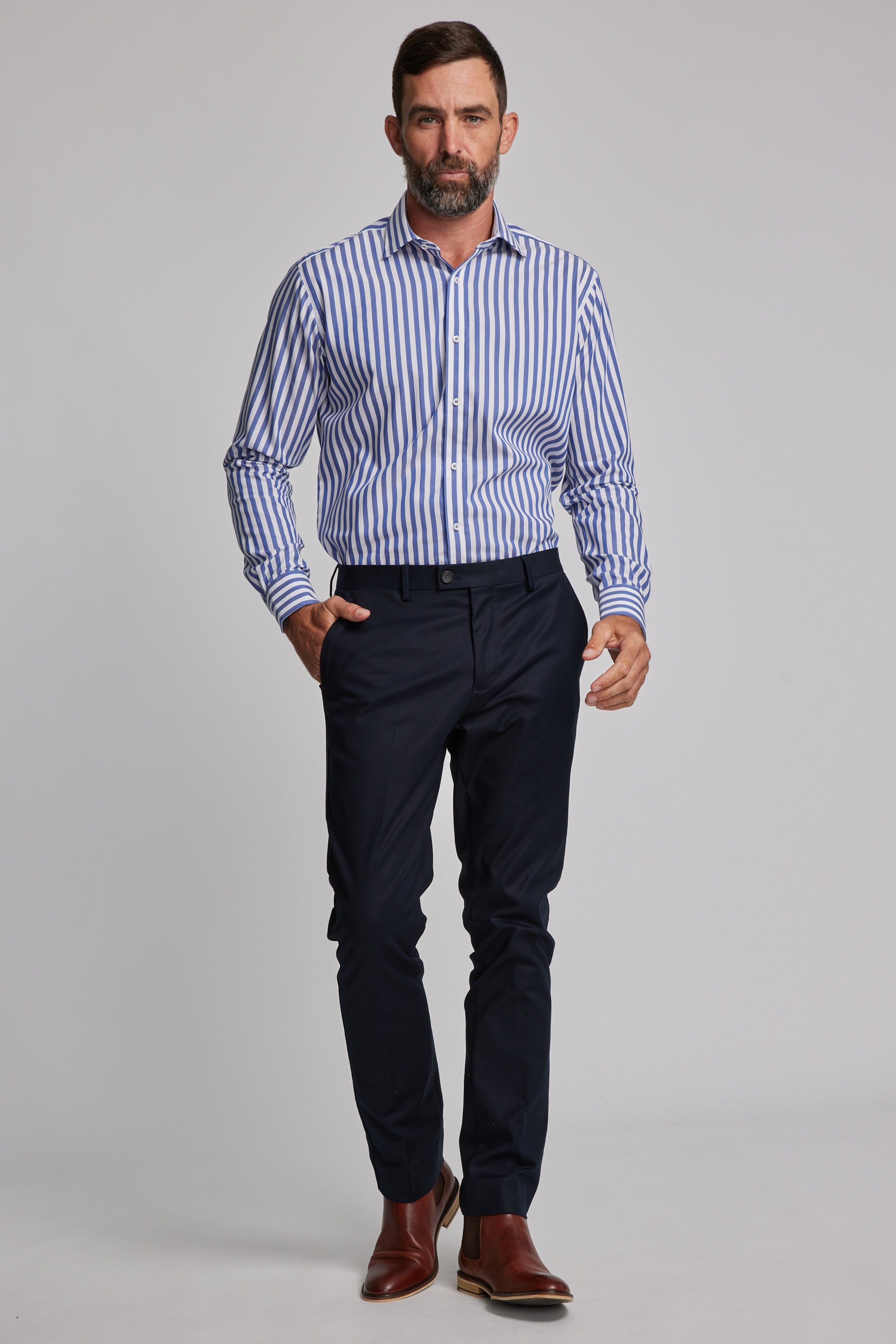 A man with short brown hair and a beard wears the Barkers Midway Shirt in a tailored fit, paired with dark navy dress pants and brown dress shoes, walking confidently against a plain light gray background.