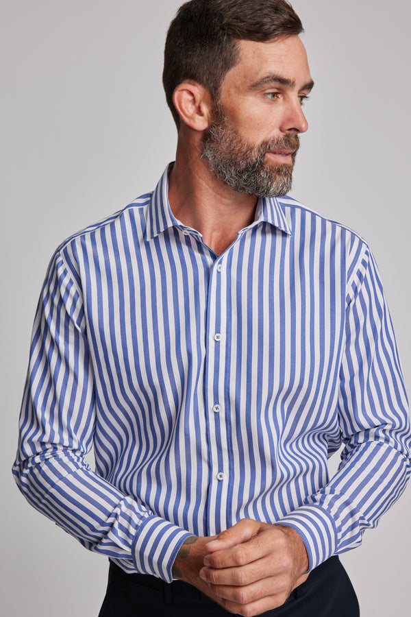 A man with short dark hair and a beard wears the Barkers Midway Shirt, a tailored fit blue striped business shirt, looking to his left with his hands clasped in front.