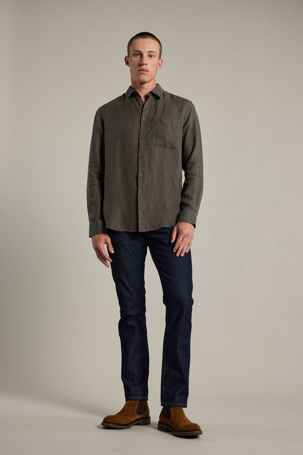 A man stands against a plain background wearing the Barkers Delano Linen Shirt in gray, paired with dark blue jeans and brown suede boots. He faces forward with a neutral expression and relaxed posture.