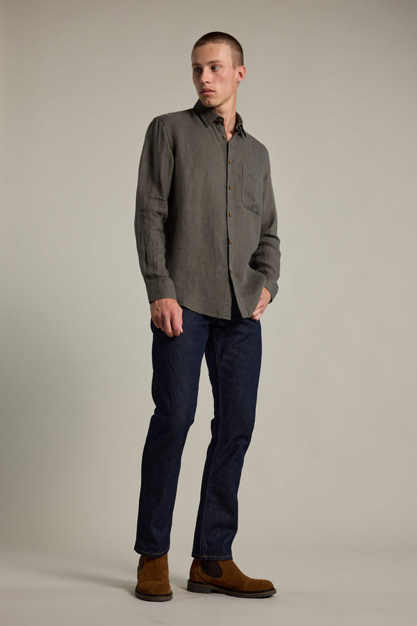 A person stands in a neutral studio wearing the Barkers Delano Linen Shirt in gray, paired with dark blue jeans and brown suede boots. They have short hair, look to the side, and keep one hand in their pocket.
