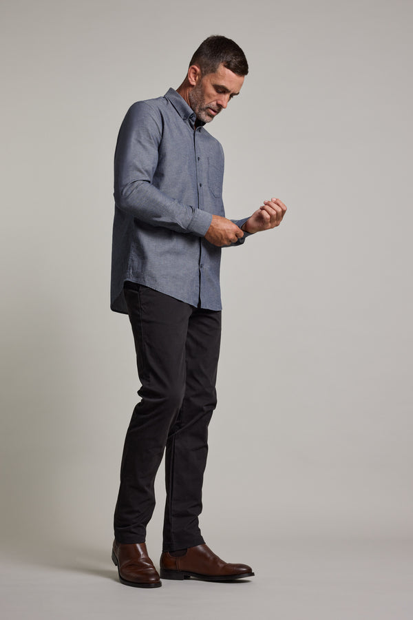 A man stands against a plain background, looking down as he fastens the cuff of his Barkers Tyson Texture Shirt. He pairs the tailored fit, long-sleeve blue shirt with black pants and brown dress shoes.