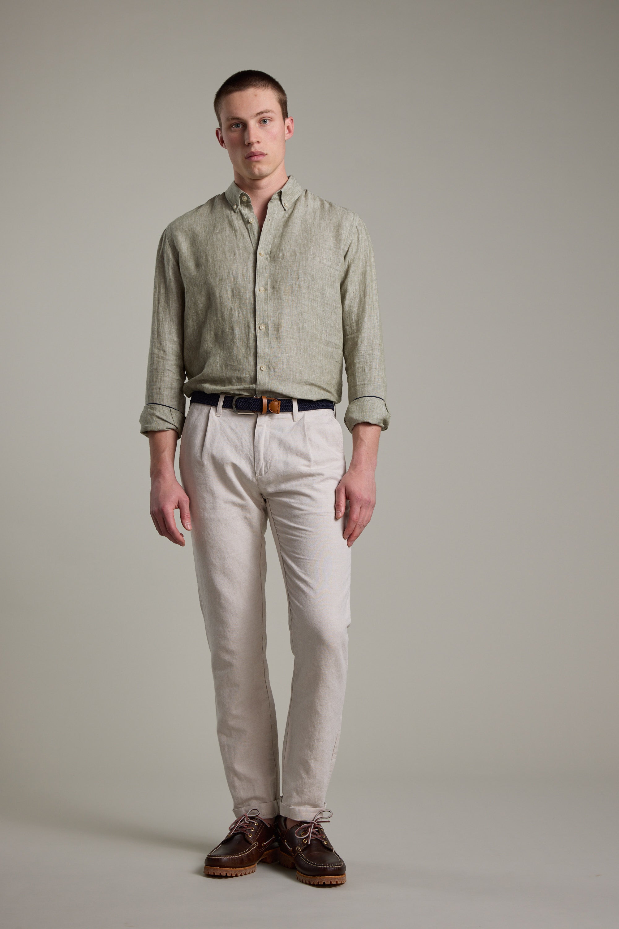 A man stands before a plain gray backdrop wearing the Barkers Dorset Linen Shirt in light green, tailored fit, paired with beige pants, a black belt, and brown shoes. His arms are by his sides and he has a neutral expression.