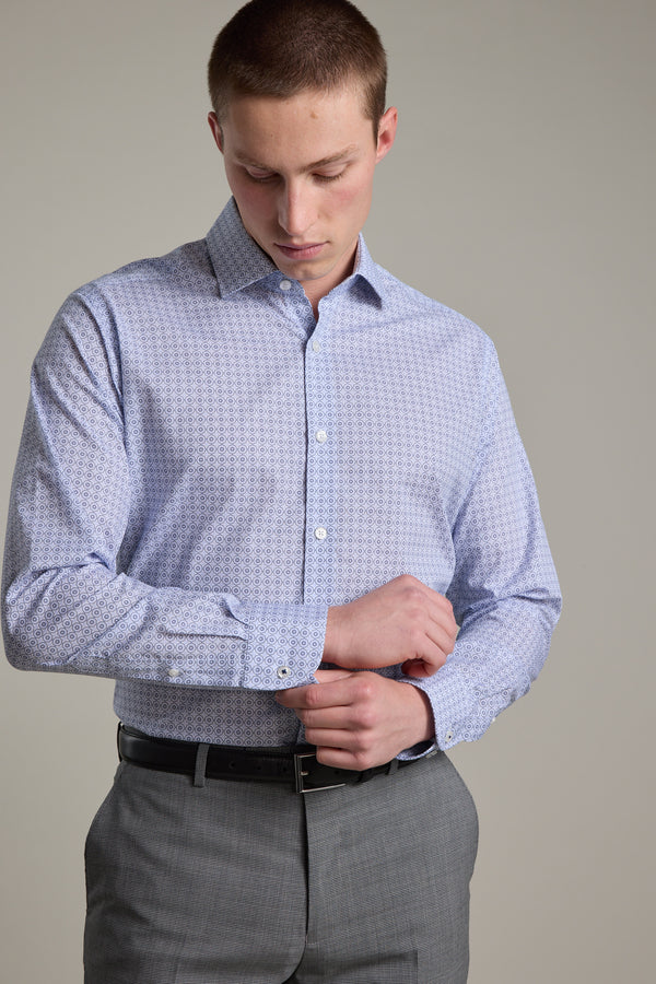 A man wearing the Barkers Seton Print Business Shirt in light blue adjusts his cuff while standing against a plain light background, paired with gray pants.