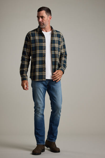 A man stands against a plain light gray background, looking to his left, wearing the Barkers Brennan Check Overshirt in green and yellow plaid over a white t-shirt, blue jeans, and brown boots.