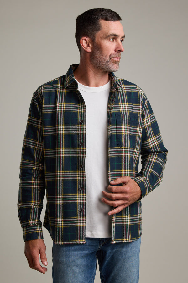 A man with short dark hair and a beard wears the Barkers Brennan Check Overshirt in green and yellow plaid over a white t-shirt and blue jeans, standing against a plain background and looking to his left.