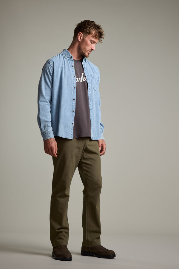 A man wears the Barkers Ruxton Denim Shirt as an overshirt over a dark tee, paired with olive green pants and dark shoes. He stands slightly sideways, looking down with relaxed hands against a neutral background.
